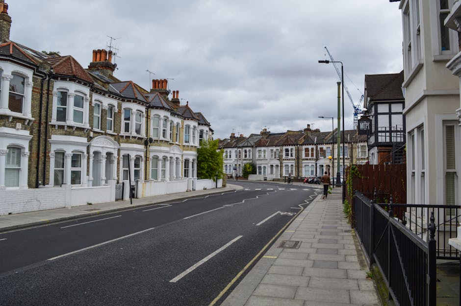 The image depicts a residential street scene on a cloudy day, with a row of Victorian-style terraced houses featuring bay windows, decorative brickwork, and chimneys, lining the left side of the street. The houses have white facades with some brickwork exposed and red tiled roofs. The pavement on the right side is made of concrete slabs with a black metal railing running along the front of several houses. A few pedestrians are visible walking along the sidewalk beneath streetlamps and lampposts, which are also present along the roadside. The road is paved with asphalt, marked with white dashed lines for parking bays and lane divisions. There are no moving vehicles currently on the road, but a crane can be seen in the background, indicating possible construction activity. This scene reflects typical urban residential moving logistics, with no visible furniture or packing materials, suggesting a quiet moment during a property relocation managed by Man with Van Little Ilford as part of their removals service.