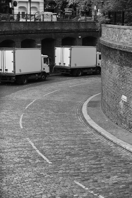 A black-and-white photograph shows two medium-sized box trucks parked on a paved, curved road in an urban area. The trucks are positioned parallel to a brick wall on the right side of the image, with the rear sections facing away from the camera. In the foreground, a cobblestone pathway curves around the brick wall, leading toward the trucks. In the background, there is a bridge or elevated structure with arches, and a railing above it, suggesting a residential or commercial district. The scene is well-lit, with visible details of the vehicles' sides and the surrounding environment. This image captures elements associated with house removals and furniture transport, illustrating delivery vehicles on a typical residential street, which could be part of a home relocation or moving process supported by Man with Van Little Ilford.