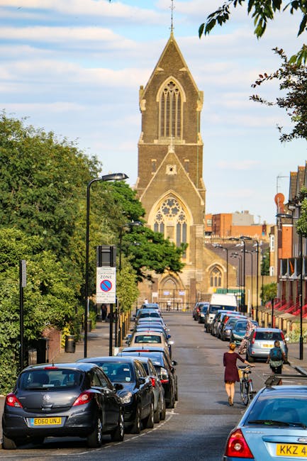A street scene in Little Ilford featuring a large stone church with a tall, pointed steeple and Gothic-style windows, flanked by leafy green trees on the left side. The street is lined with parked cars on both sides, including compact and mid-sized vehicles, with some showing visible license plates. In the foreground, a woman wearing a burgundy coat is walking while pushing a bicycle on the sidewalk, and another person nearby is also walking. The scene includes street lamps, signage, and a clear sky with scattered clouds overhead. The image captures the urban environment during daytime, illustrating the typical setting where home relocation or furniture transport services by Man with Van Little Ilford may take place, especially during convenient, daylight moving activities.