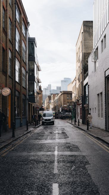 A narrow urban street scene during daytime, with commercial and residential buildings on both sides. The buildings feature varying architectural styles, including brick facades and modern white exteriors. A white van is parked on the left side, partially blocking the pavement, which is lined with black bollards. The street surface appears wet, possibly after recent rain, with visible puddles and reflective patches. In the background, taller modern skyscrapers are visible under a cloudy sky. The scene illustrates a typical city environment suitable for local house removals or furniture transport services, with a focus on the logistics of navigating tight urban spaces, as undertaken by companies like Man with Van Little Ilford during home relocation and packing and moving operations.