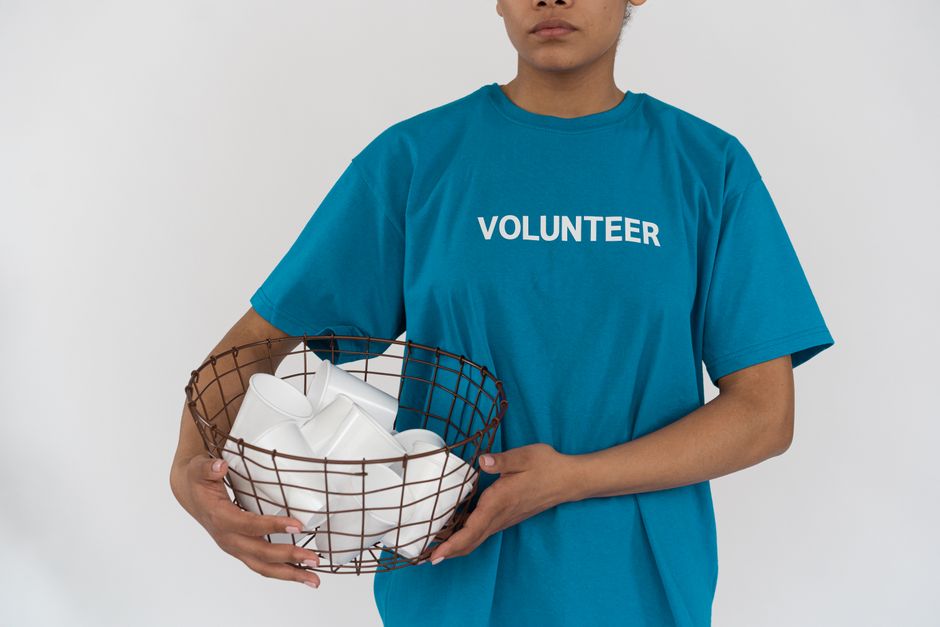 A person wearing a bright blue T-shirt with the word 'VOLUNTEER' printed in white across the chest is holding a round wire basket filled with crumpled white packing paper or protective padding. The individual is standing indoors against a plain white background, with only their torso and part of their arms visible. The basket is being held with both hands, one supporting the bottom and the other gripping the side, suggesting a careful handling process typical of moving or packing activities. This scene may depict a volunteer assisting with packing or preparing items for a house removal or relocation service, such as those offered by Man with Van Little Ilford, supporting efficient and eco-friendly packing methods during the furniture transport or home relocation process.