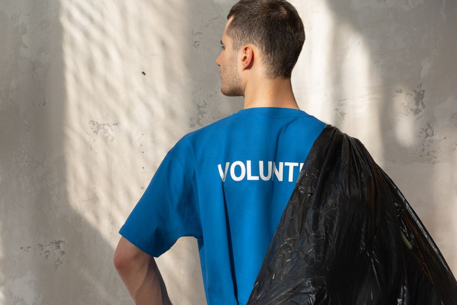 A male volunteer wearing a bright blue T-shirt with the word 'VOLUNTEER' printed on the back stands outdoors against a textured, light grey wall. He has short, dark hair and is holding a large black plastic trash bag over his shoulder, filled with packing materials or items to be discarded. The scene is illuminated by natural daylight, and the volunteer appears to be involved in a home relocation or cleaning activity as part of a furniture transport or packing and moving process. The photograph emphasizes the volunteer’s role in assisting with removals, consistent with the services offered by Man with Van Little Ilford in the context of house removals and eco-friendly relocation efforts.