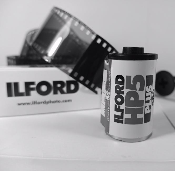 A black and white photograph showing a roll of 35mm photographic film partially unspooled, with the film extending from the roll and draped over a white cardboard box. The box has the text 'ILFORD' printed on its side, along with a website link. Behind the box, there is an open film canister with a segment of film strip visible, featuring perforations along its edges. The scene is set on a flat surface, likely a table, with a plain background. The image depicts elements related to film storage and processing, relevant to the process of packing and transporting photographic equipment during a home relocation or moving service offered by Man with Van Little Ilford.