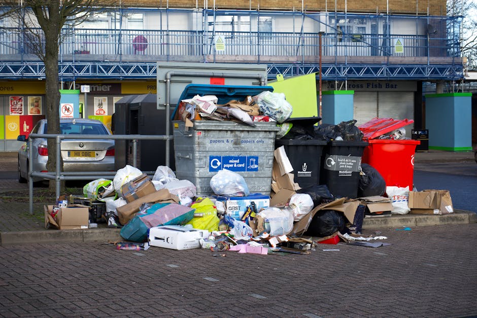 A collection of overflowing waste and recycling bins situated on a paved pavement in a suburban area, with various waste materials including cardboard boxes, plastic bags, paper, and packaging strewn 