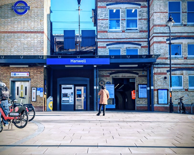 A woman wearing a beige coat and black leggings stands outside the entrance of Hanwell station, a brick building with large blue signage indicating the station name. The station features a modern blac