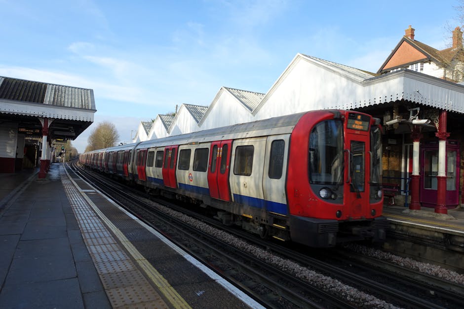 A woman wearing a beige coat and black leggings stands outside the entrance of Hanwell station, a brick building with large blue signage indicating the station name. The station features a modern blac