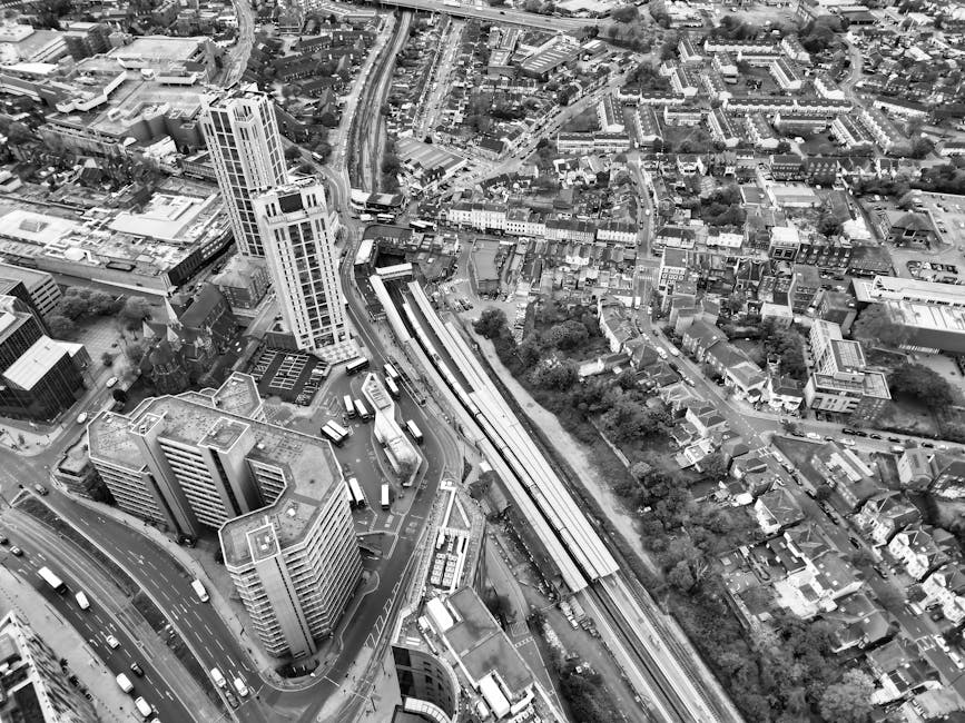 Aerial black-and-white photograph of a dense urban area featuring high-rise office buildings and residential structures, with a railway station and train tracks running parallel, adjacent to a busy mu