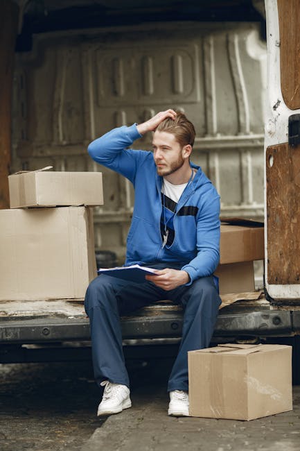 A man wearing a white t-shirt, blue jeans, a black cap, and white sneakers is standing outside on the pavement, carrying three large cardboard boxes stacked one on top of the other. The boxes are held