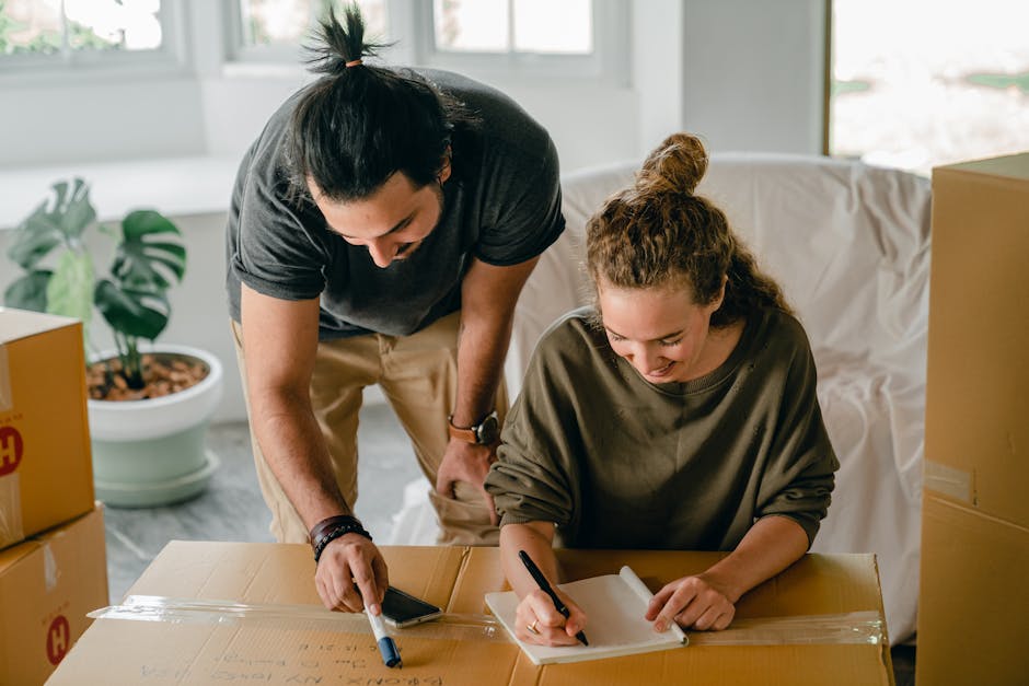 A man and woman are inside a room during a house move, surrounded by cardboard boxes. The man is leaning over a large cardboard box, using a marker to label it, while the woman is seated at the table, also writing on a smaller cardboard box or a note. The woman has curly hair tied back and is wearing a dark green long-sleeve top, and the man has dark hair pulled back in a bun, wearing a black t-shirt and beige pants. There are packing materials, such as plastic wrap and tape, visible on the boxes, and a potted plant and a white sofa are seen in the background, indicating a home environment. The space is well-lit with natural light coming through windows, and the scene depicts a typical packing and loading process as part of home relocation, suitable for a house removals service like Man with Van Little Ilford.