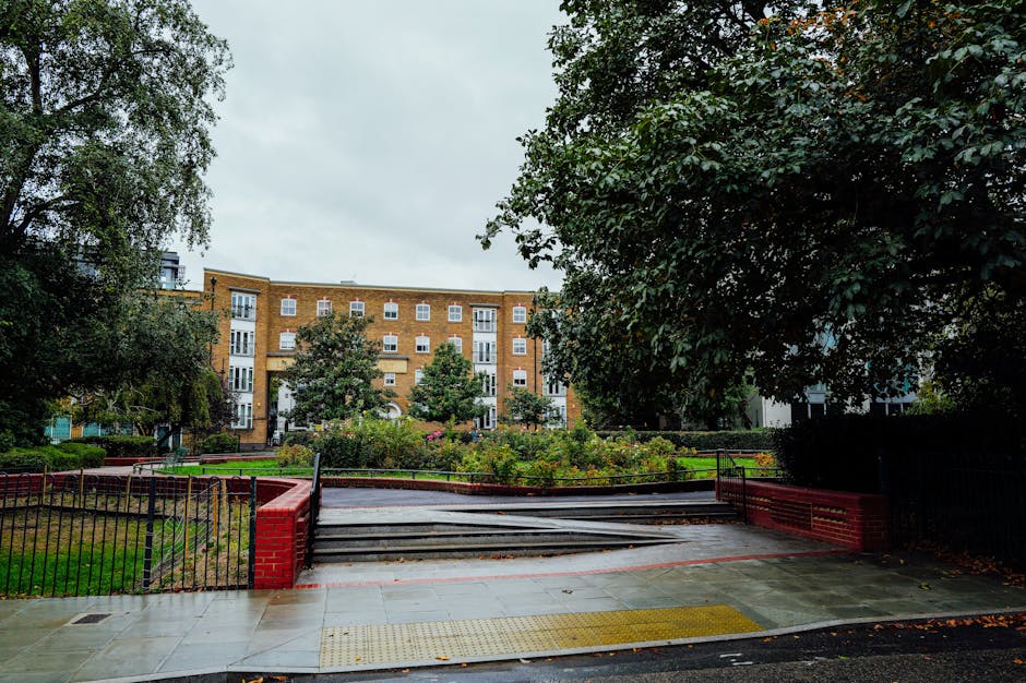 A view of a residential outdoor area showing a paved walkway leading to a set of short stairs with metal railings on both sides, positioned in front of a mid-rise brick apartment building. The stairs appear wet, indicating recent rain, and are surrounded by greenery including trees, shrubs, and flowerbeds. To the left, a green lawn is bordered by a black metal fence with a red brick pillar at the corner. Large leafy trees frame the scene on both sides, casting partial shade over the area. The multi-storey apartment building in the background has a beige brick facade with numerous white-framed windows and small balconies. The sky above is overcast with grey clouds, suggesting typical weather conditions in Ilford. This setting, with its accessible pathway and entryway, is relevant to house relocations and furniture transport planning by Man with Van Little Ilford, highlighting potential access points and environmental considerations during home relocation or removal services.