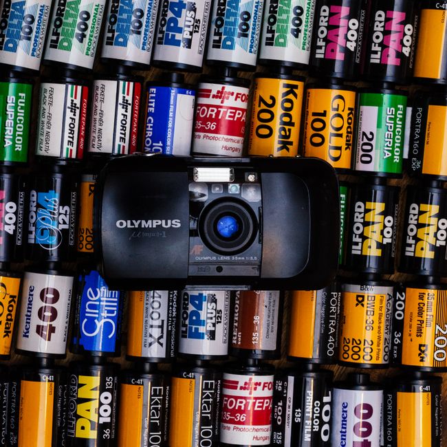 A close-up aerial view showing an assortment of photographic film rolls arranged on a flat surface, predominantly black, white, yellow, and green in colour, with some labels displaying brand names such as Kodak, Ilford, and Fujifilm along with film speed ratings like 200, 400, and 135-36. In the center of the image, there is a black Olympus compact film camera positioned among the rolls, with the lens facing slightly upward and the camera's branding clearly visible. The surrounding film rolls are tightly packed, with some standing vertically and others lying horizontally, creating a textured, grid-like pattern. The lighting is even, highlighting the variety of colours and the details on the film roll labels, which suggests an indoor environment used for organised storage of photographic materials, relevant for contexts involving packing and transport during home or flat relocations, such as those managed by Man with Van Little Ilford.